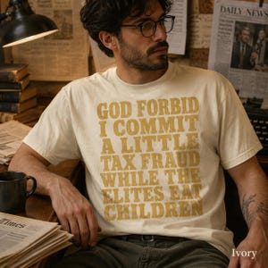 Op de afbeelding: Een ivoorkleurig T-shirt met korte mouwen en opvallende, verweerde mosterdgele tekst die luidt: GOD FORBID I COMMIT A LITTLE TAX FRAUD WHILE THE ELITES EAT CHILDREN. Het T-shirt heeft een ronde hals en een standaard pasvorm.