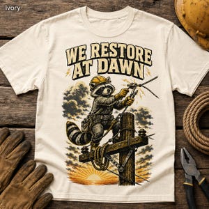 May include: An ivory short-sleeve t-shirt featuring a graphic of a raccoon wearing a yellow hard hat and utility belt, standing on a utility pole. The text "WE RESTORE AT DAWN" is printed above the raccoon, which is holding electrical wires.