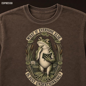 May include: A dark brown crew neck t-shirt featuring a graphic illustration of a frog holding a book with one hand on its chin. The text reads "WHAT IF EVERYONE ELSE IS JUST UNDERTHINKING?" surrounded by a decorative border of reeds and lily pads.