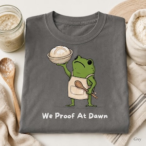 Könnte beinhalten: Ein anthrazitfarbenes T-Shirt mit einer Illustration eines grünen Frosches in einer beigen Schürze, der einen Holzlöffel und einen runden Laib Brot in einem Gärkörbchen hält, mit dem weißen Text "We Proof At Dawn" unter dem Motiv.