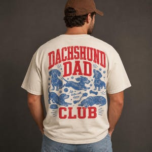 Peut inclure: Un t-shirt &agrave; manches courtes blanc cass&eacute; avec un motif rouge et bleu au dos. Le texte indique "DACHSHUND DAD CLUB" et "life is better with doxies", accompagn&eacute; d'illustrations de quatre teckels dans diverses positions avec des jouets.