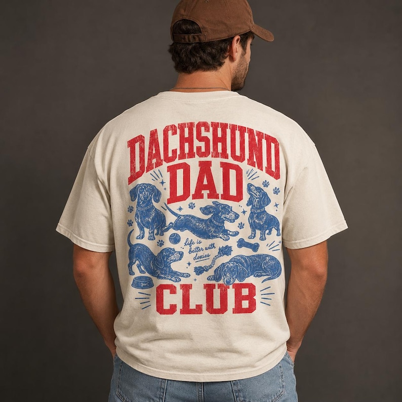Peut inclure: Un t-shirt &agrave; manches courtes blanc cass&eacute; avec un motif rouge et bleu au dos. Le texte indique "DACHSHUND DAD CLUB" et "life is better with doxies", accompagn&eacute; d'illustrations de quatre teckels dans diverses positions avec des jouets.