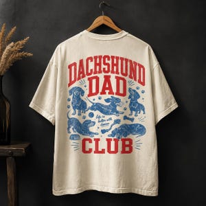 Peut inclure: Un t-shirt beige &agrave; manches courtes avec un motif au dos portant le texte rouge DACHSHUND DAD CLUB. Le design comprend des illustrations bleues de teckels dans diverses positions, une balle, un os et le texte life is better with doxies.