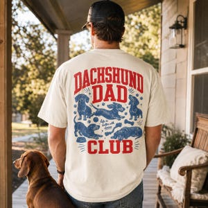 Peut inclure: Un t-shirt &agrave; manches courtes de couleur cr&egrave;me avec un motif rouge et bleu au dos. Le texte indique DACHSHUND DAD CLUB et Life is better with doxies. Le design comprend des illustrations de quatre teckels, une balle, un os et des empreintes.