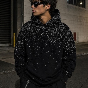 May include: Black hoodie with a constellation-like pattern of small, white embellishments. The hoodie has a drawstring hood and a front pocket. The person in the image is wearing black sunglasses and black pants.