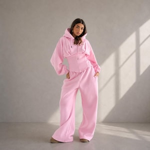Puede incluir: Un conjunto de dos piezas de color rosa claro que consta de una sudadera con capucha con un corpiño estilo corsé y pantalones de chándal de pernera ancha. La sudadera tiene mangas largas y una capucha con cordón.