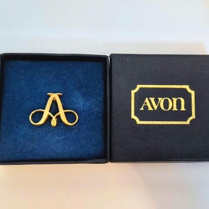 May include: A gold-toned metal brooch shaped like the letter A, resting inside a square black box with a blue velvet lining. The box lid is black with the word Avon printed in gold lettering inside a decorative gold-bordered rectangular frame.