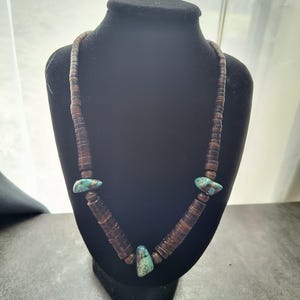 Op de afbeelding: Een bruine kralenketting met drie turkooizen stenen. De ketting wordt getoond op een zwarte mannequin. De kralen zijn plat en cirkelvormig en de turkooizen stenen zijn onregelmatig van vorm.