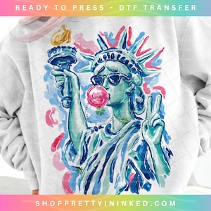 May include: White sweatshirt featuring a colorful graphic of the Statue of Liberty wearing sunglasses and blowing a bubblegum bubble. The design includes the text "READY TO PRESS - DTF TRANSFER" and "SHOPPRETTYININKED.COM".