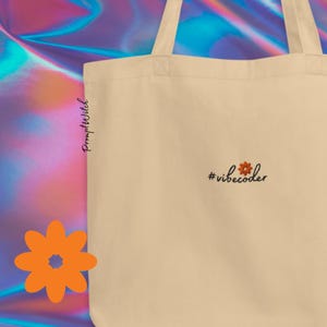 Puede incluir: Un bolso tote de lona beige con el texto bordado en negro #vibecoder, donde un pequeño icono de flor naranja reemplaza el punto de la i. El bolso tiene dos asas para el hombro y una forma rectangular sencilla, ofreciendo una base neutra.