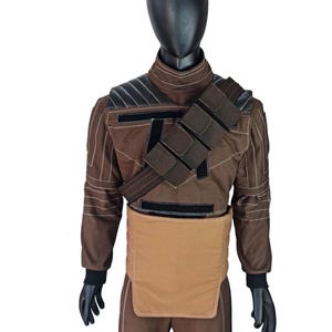 Puede incluir: Traje marr&oacute;n con detalles en negro, exhibido en un maniqu&iacute;. El atuendo presenta una chaqueta con hombreras, una correa en el pecho con bolsillos y una pieza beige tipo delantal alrededor de la cintura. Guantes negros y cabeza negra completan el conjunto.