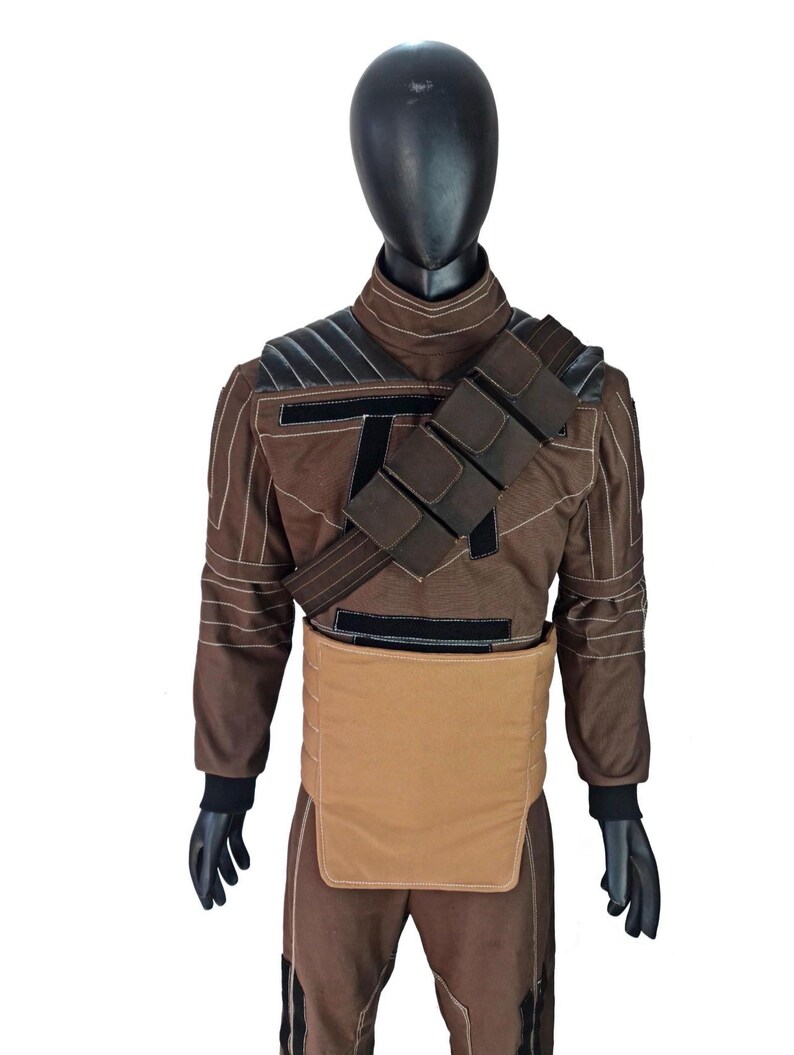 Puede incluir: Traje marr&oacute;n con detalles en negro, exhibido en un maniqu&iacute;. El atuendo presenta una chaqueta con hombreras, una correa en el pecho con bolsillos y una pieza beige tipo delantal alrededor de la cintura. Guantes negros y cabeza negra completan el conjunto.