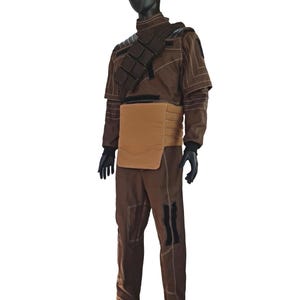 Puede incluir: Un disfraz marr&oacute;n exhibido en un maniqu&iacute;. El atuendo incluye una chaqueta de cuello alto con costuras blancas, una pieza para el pecho y pantalones a juego. Guantes y botas negras completan el conjunto. El disfraz tiene una est&eacute;tica futurista o militar.