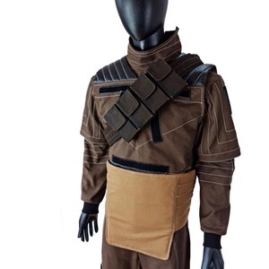 Puede incluir: Un disfraz marr&oacute;n exhibido en un maniqu&iacute;. El disfraz presenta una chaqueta marr&oacute;n con detalles negros, una correa para el hombro con bolsillos y una pieza pectoral beige. El maniqu&iacute; lleva guantes negros y una cabeza negra.