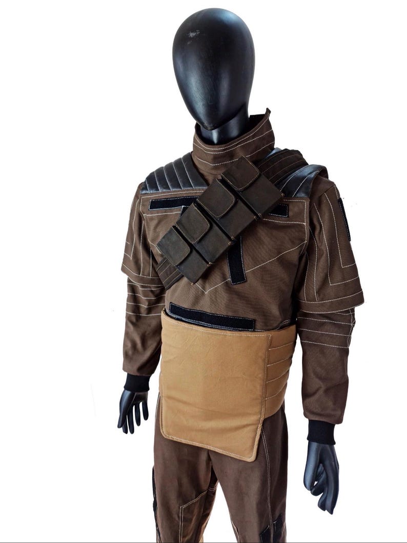 Puede incluir: Un disfraz marr&oacute;n exhibido en un maniqu&iacute;. El disfraz presenta una chaqueta marr&oacute;n con detalles negros, una correa para el hombro con bolsillos y una pieza pectoral beige. El maniqu&iacute; lleva guantes negros y una cabeza negra.