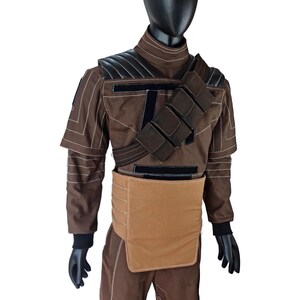 Puede incluir: Traje marr&oacute;n con detalles en negro, exhibido en un maniqu&iacute;. El atuendo presenta un cuello alto, hombreras y una correa en el pecho con bolsillos. Los pantalones tienen parches negros en las rodillas. Guantes y cabeza negros completan el look.