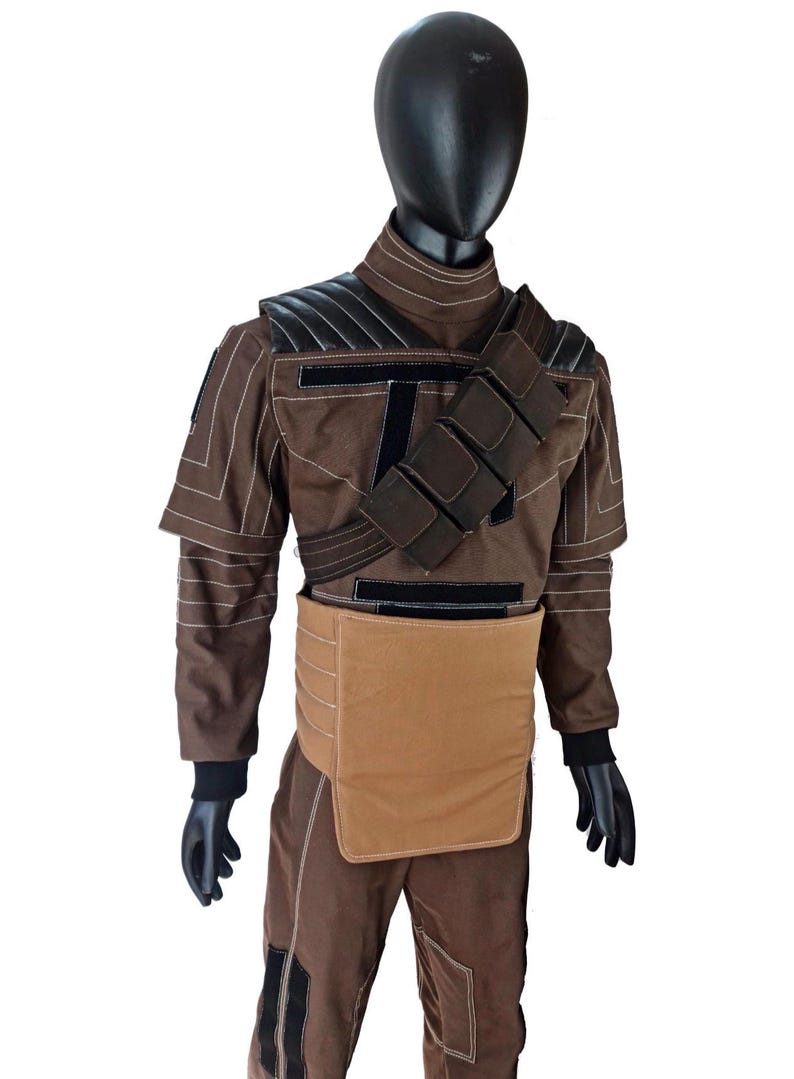 Puede incluir: Traje marr&oacute;n con detalles en negro, exhibido en un maniqu&iacute;. El atuendo presenta un cuello alto, hombreras y una correa en el pecho con bolsillos. Los pantalones tienen parches negros en las rodillas. Guantes y cabeza negros completan el look.