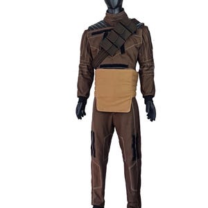 Puede incluir: Mono marr&oacute;n con protector pectoral beige y arn&eacute;s negro. El atuendo tiene detalles en negro y costuras blancas. El maniqu&iacute; lleva guantes negros y botas negras. El atuendo se exhibe en un maniqu&iacute; negro.