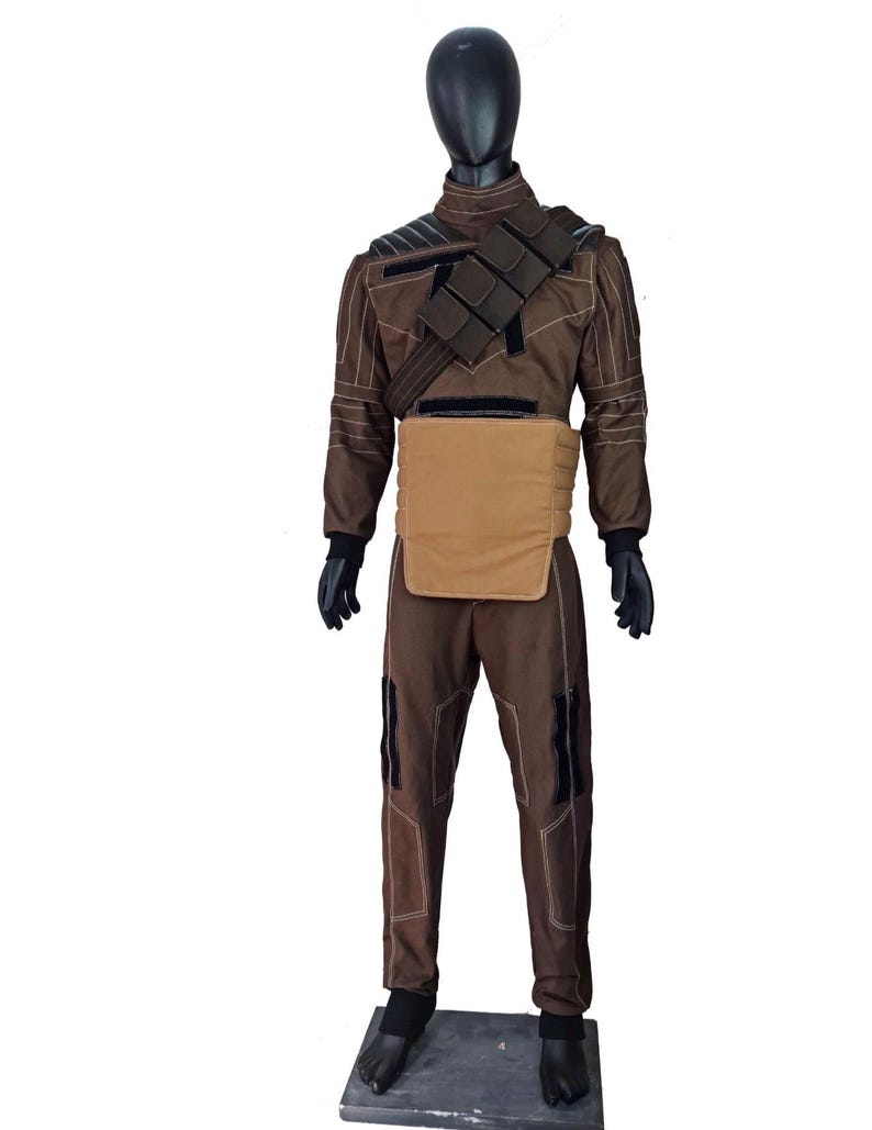 Puede incluir: Mono marr&oacute;n con protector pectoral beige y arn&eacute;s negro. El atuendo tiene detalles en negro y costuras blancas. El maniqu&iacute; lleva guantes negros y botas negras. El atuendo se exhibe en un maniqu&iacute; negro.