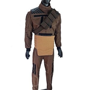 Puede incluir: Traje marr&oacute;n con detalles negros, exhibido en un maniqu&iacute;. El atuendo incluye una chaqueta de manga larga, pantalones y una pieza para el pecho. La chaqueta tiene un cuello alto y acolchado en los hombros. El disfraz tambi&eacute;n presenta un cintur&oacute;n con bolsas y guantes negros.