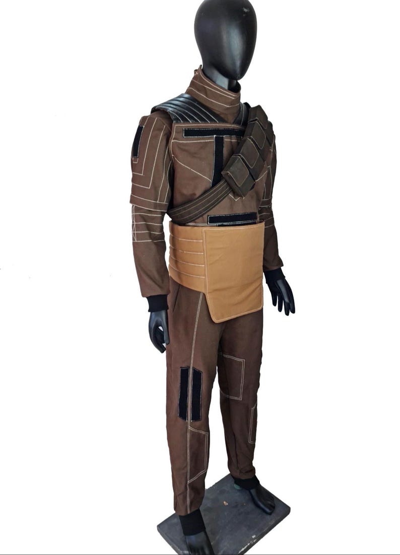 Puede incluir: Un disfraz marr&oacute;n con detalles en negro, exhibido en un maniqu&iacute;. El atuendo incluye una chaqueta con protecci&oacute;n para los hombros, pantalones y un cintur&oacute;n con una bolsa. El disfraz tiene costuras blancas y guantes negros.