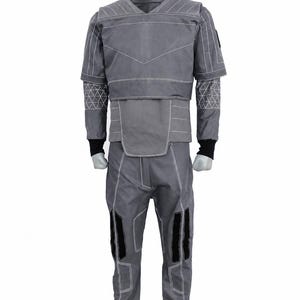Puede incluir: Mono gris con costuras blancas y detalles negros. El traje presenta un cuello alto, mangas cortas y patrones geom&eacute;tricos en los brazos. Los pantalones tienen rodilleras negras. El dise&ntilde;o sugiere un tema futurista o t&aacute;ctico.