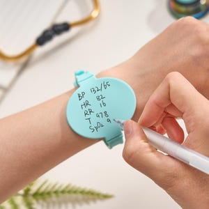 May include: A light blue silicone wristband featuring a circular writing surface for notes. The surface displays handwritten medical vitals including BP 132/65, HR 82, RR 16, T 97.8, and SpO2 9. The band is secured around the wrist with a clasp mechanism.