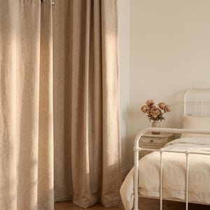 Peut inclure: Rideaux beiges allant jusqu'au sol avec une texture tissée visible, dotés d'œillets métalliques argentés suspendus à une tringle à rideaux argentée assortie. Les rideaux pendent en plis verticaux, couvrant un coin de la pièce.