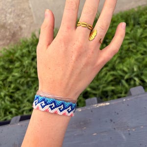 May include: A gold ring with a leaf design and a gold ring with a woven design are on a hand. A woven friendship bracelet in shades of blue, pink, and white is on the wrist. The bracelet has a geometric pattern.