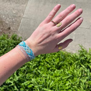May include: A wrist adorned with a blue, white, and gray woven bracelet, complemented by delicate pink string bracelets. Two gold rings with fan-shaped designs and a snake-like ring add a touch of elegance. The hand is set against a backdrop of green foliage.