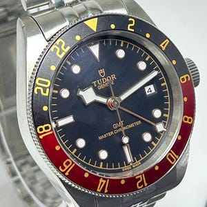 May include: A stainless steel Tudor GMT wristwatch with a black dial, luminous hour markers, and a date window at the three o'clock position. The bidirectional rotating bezel features a black and red insert with gold-colored 24-hour numerals.