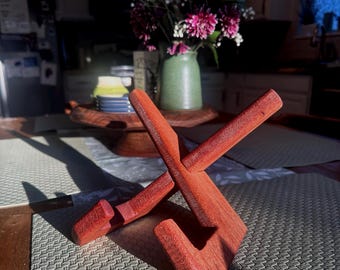 May include: A reddish-brown wooden display stand composed of two interlocking pieces that form an X-shaped structure. The stand features two notched arms designed to hold an object, with a smooth, natural wood grain finish visible throughout the piece.