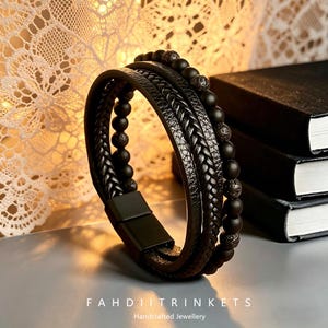 May include: A multi-layered black bracelet with braided and beaded elements. The bracelet features a magnetic clasp and is displayed on a light gray surface. The text "FAHDII TRINKETS Handcrafted Jewellery" is visible at the bottom.