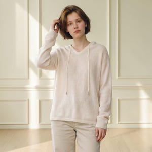 May include: A long-sleeved, cream-colored hooded sweater featuring a V-neckline and matching drawstring cords with metallic tips. The garment is crafted from a lightweight, fine-knit fabric and is paired with light-colored trousers.