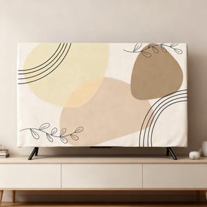 Pode incluir: Uma capa retangular para TV com um design abstrato. A capa tem um fundo creme com formas suaves em tons de bege e castanho claro. Linhas pretas curvas e detalhes de folhas adicionam contraste. A capa é colocada sobre uma TV de tela plana.