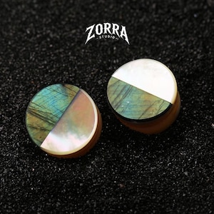 Puede incluir: Un par de expansores de oreja circulares con un diseño dividido, con una mitad hecha de labradorita verde iridiscente y la otra mitad de nácar blanco. El texto ZORRA STUDIO está impreso en blanco sobre las piezas de joyería.