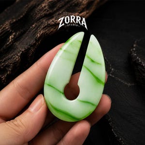 May include: A pale green ear weight with a smooth, polished finish and dark green veining, featuring a teardrop shape with a central circular cutout and a top opening. The Zorra Studio logo is printed in white above the jewelry piece.