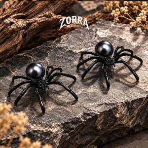 Puede incluir: Dos broches con forma de araña que presentan patas de metal negro brillante y un cuerpo de perla grande, redonda e iridiscente de color gris oscuro. El texto ZORRA STUDIO es visible sobre las arañas. Los broches están sobre una piedra gris.