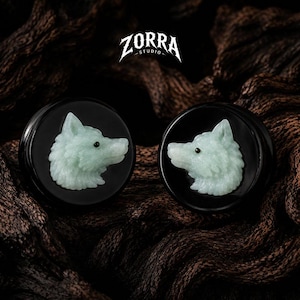 Puede incluir: Un par de expansores circulares negros con una cabeza de lobo tallada en color verde pálido en cada cara. Las tallas de lobo tienen pequeños ojos negros y están sobre un fondo oscuro y liso. El texto ZORRA STUDIO es visible en la parte superior.