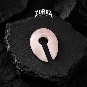 Pode incluir: Um alargador de orelha oval rosa claro com um design de buraco de fechadura. O alargador é feito de um material translúcido e assenta numa superfície escura e texturizada. O logótipo ZORRA STUDIO é visível na parte superior da imagem.