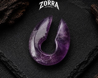 Puede incluir: Un peso para oreja de amatista morada pulida con un diseño en forma de lágrima y una abertura ancha en la parte superior. La piedra muestra inclusiones naturales y un acabado liso y brillante. El texto ZORRA STUDIO aparece impreso arriba.