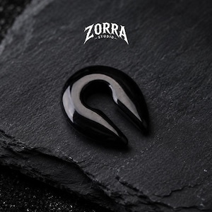 May include: A glossy black ear gauge, shaped like a teardrop, sits on a dark, textured surface. The gauge has a smooth, curved design with a hollow center. The ZORRA STUDIO logo is visible in the upper portion of the image.