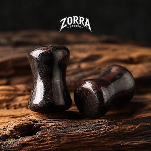 May include: Two dark red and black stone ear plugs with a glossy finish. The plugs are on a wooden surface. The logo "ZORRA STUDIO" is at the top of the image.
