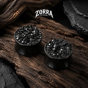 Puede incluir: Un par de dilatadores de oreja negros con una superficie texturizada y cristalina de aspecto facetado. El texto ZORRA STUDIO es visible sobre los artículos. Los dilatadores tienen un cuerpo cilíndrico liso y una superficie superior brillante.