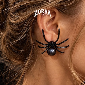 Puede incluir: Un pendiente en forma de araña negra con una perla gris oscuro brillante como abdomen. La araña tiene ocho patas delgadas que se curvan alrededor del lóbulo. El texto ZORRA STUDIO es visible en letras blancas sobre la oreja.