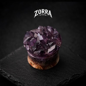 May include: A purple amethyst crystal plug with a natural stone base. The top of the plug is a cluster of dark purple crystals. The ZORRA STUDIO logo is at the top of the image.