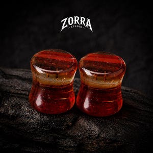 Op de afbeelding: Twee rood-bruine glazen oordopjes met een uitlopende vorm. De oordopjes hebben een gestreept patroon en staan tegen een donkere achtergrond. Het logo "ZORRA STUDIO" staat bovenaan.