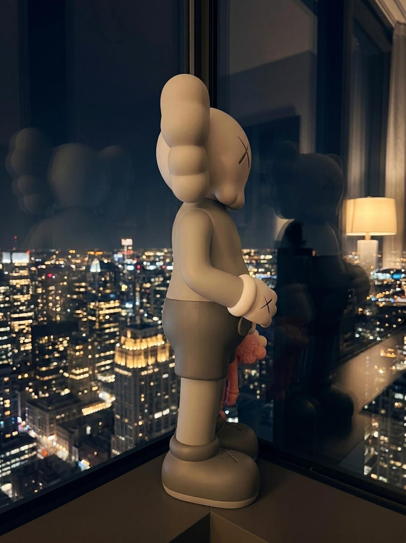 K&ouml;nnte beinhalten: Eine graue KAWS-Figur im Profil, mit einem abgerundeten Kopf mit X-f&ouml;rmigen Augen, gro&szlig;en cartoonartigen H&auml;nden mit wei&szlig;en Manschetten und einer dunkelgrauen Shorts. Die Figur hat eine glatte, matte Oberfl&auml;che vor einer Fensterscheibe.