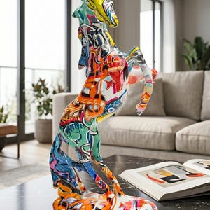 Peut inclure: Une sculpture abstraite et colorée d'un cheval cabré. La statue est recouverte d'art de style graffiti aux teintes vives d'orange, de bleu, de jaune et de vert. Elle est posée sur une table en marbre noir, à côté d'un livre ouvert.