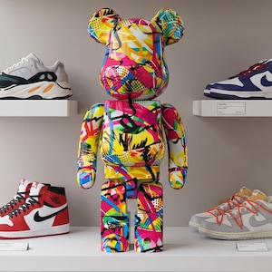 Pode incluir: Uma figura colecionável em forma de urso com um padrão vibrante de estilo graffiti multicolorido, com salpicos de rosa, amarelo, azul e preto, exibida numa prateleira rodeada por vários ténis desportivos em estantes brancas.